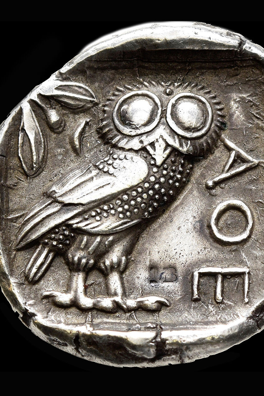 Das Bild zeigt eine antike Silbermünze mit der Darstellung einer Eule. Das Foto verlinkt auf eine Unterseite, auf der diese antike Münzen und ihr Motiv, aber auch die modernen griechischen Euro-Münzen erklärt werden.